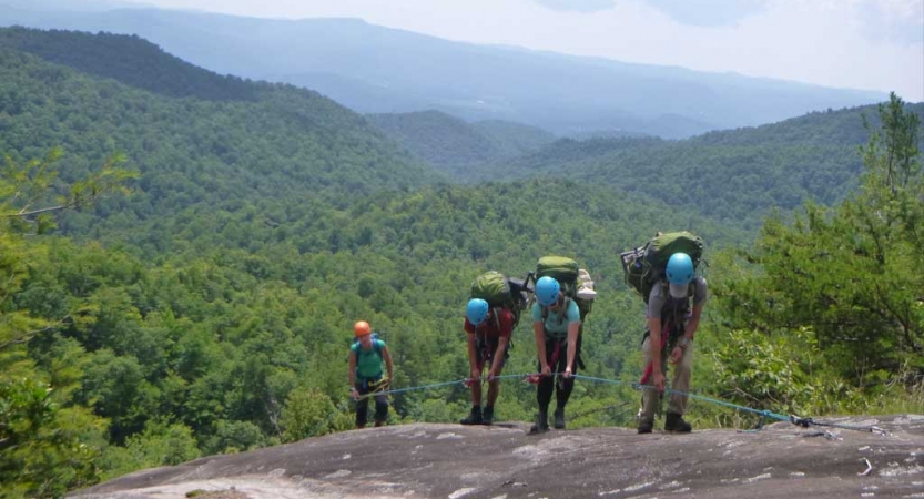 A group of students wearing helmets and backpacks hold on to a rope as they scramble up a rocky slope. Behind them is a green mountainous landscape. 